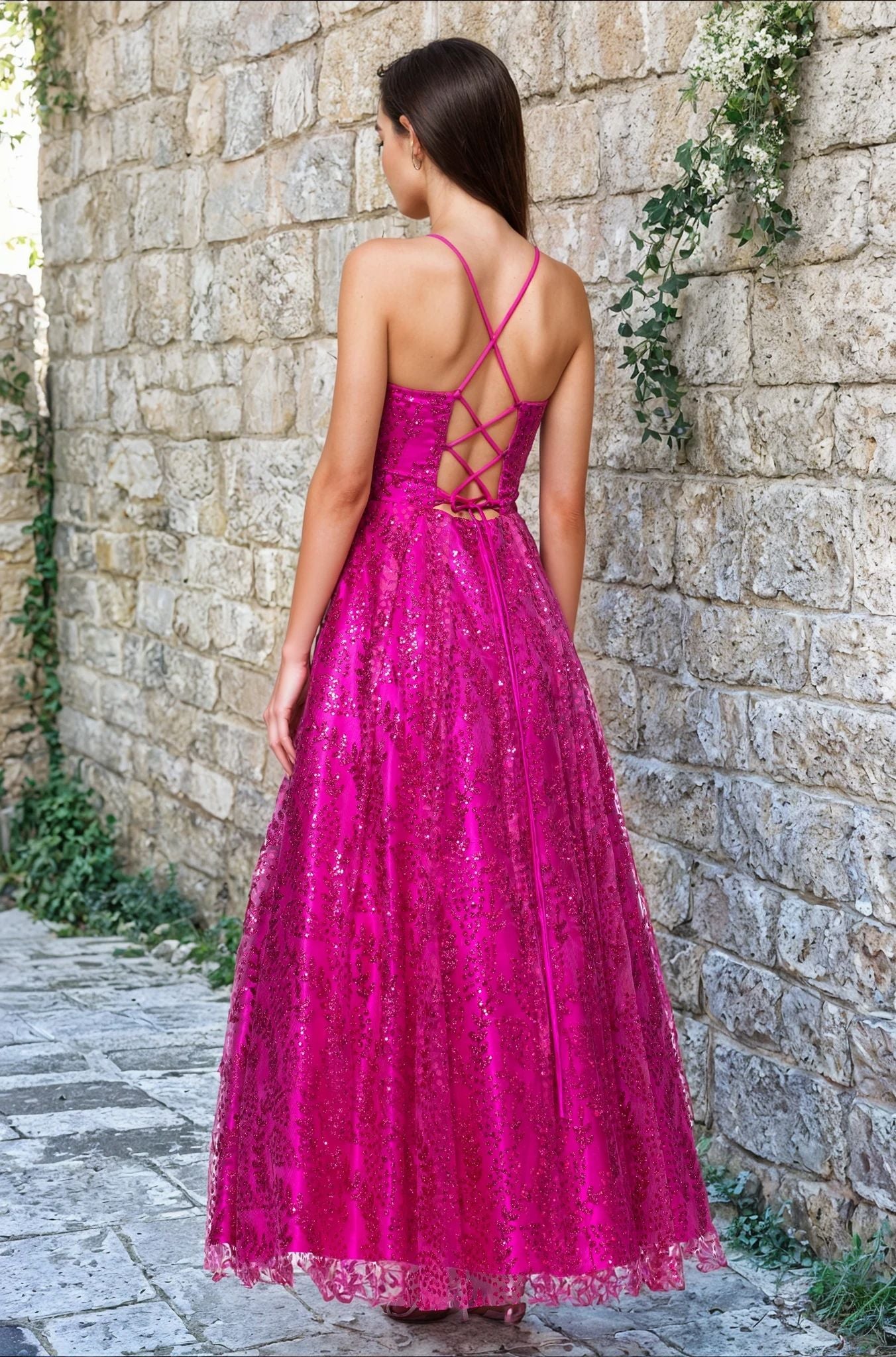 A woman stands on a stone path facing a stone wall, wearing the Blondie Nites 4867BN Long Glitter Prom Dress—a bright pink, floor-length junior formal dress with a crisscross lace-up back and shimmering glitter-print details.